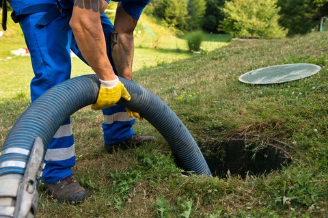 man standing over a septic tank access point using a large black hose pipe to clean the tank.